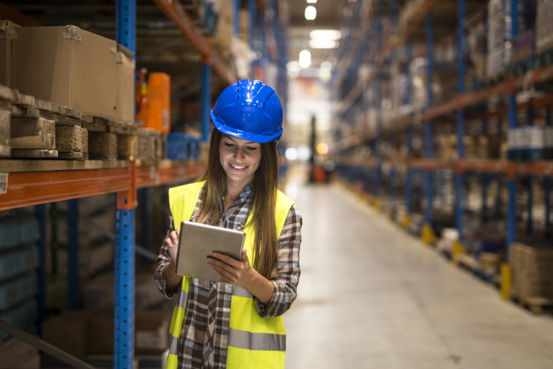 Warehouse female worker checking inventory on digital tablet in large distribution warehouse storage area.