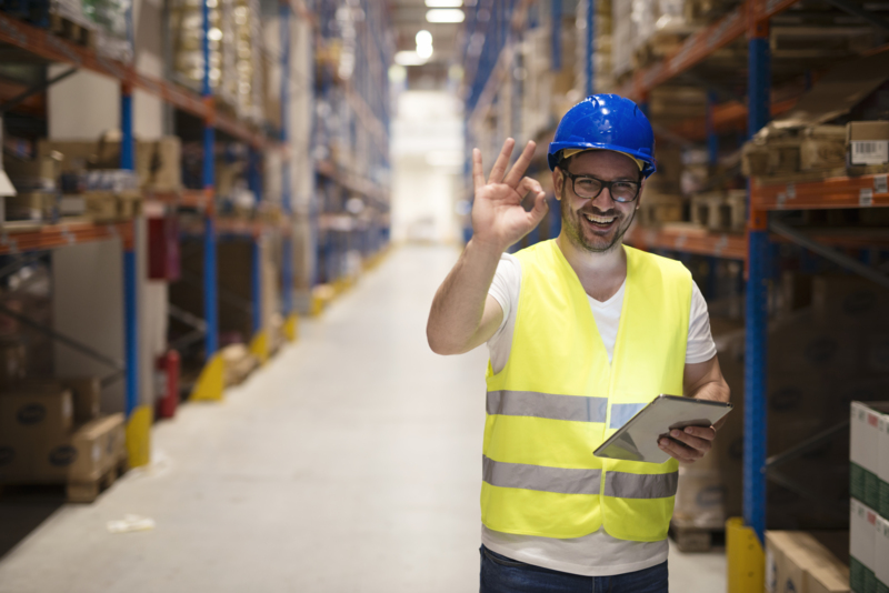 Warehouse worker standing in large storage center and showing OK hand gesture satisfied on delivering goods.